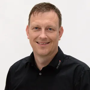 Ein Mann mit kurzen braunen Haaren, im schwarzen Arbeitshemd, lacht vor weißem Hintergrund - Thema Arbeitsschutz.
