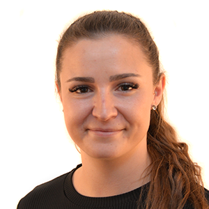 Geena Martinelli mit zurückgebundenem braunen Haar und schwarzer PSA-Oberbekleidung lächelt vor weissem Hintergrund in die Kamera.