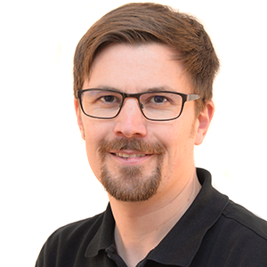 Thomas Greter, Experte für Arbeitsschutz, trägt Schutzbrille und Bart, lächelt im schwarzen Hemd vor weißem Hintergrund.