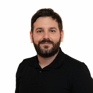 Ein Mann mit kurzem dunklem Haar und Bart trägt ein schwarzes Hemd, lächelt vor weißem Hintergrund. Arbeitsschutz Experte.