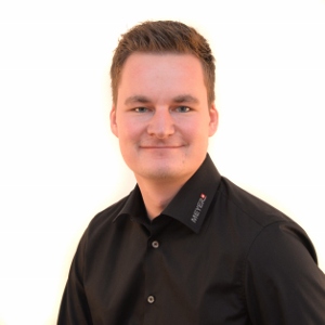 Junger Mann mit kurzem braunem Haar trägt Hemd mit Logo, posiert vor weissem Hintergrund, lächelt, Arbeitsschutz Bekleidung.