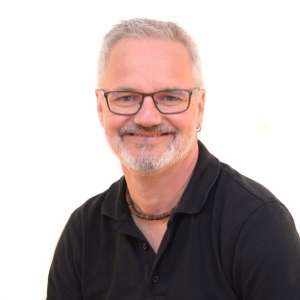 Ein Mann mit grauen Haaren, Bart und Schutzbrille lacht, trägt Poloshirt, Kette vor neutralem Hintergrund. Arbeitsschutz Experte.