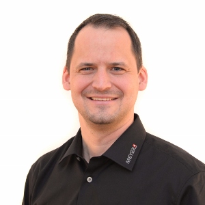 Ein Mann mit kurzen dunklen Haaren trägt ein schwarzes Poloshirt mit Logo, steht vor weißem Hintergrund, Arbeitsschutz-Experte.