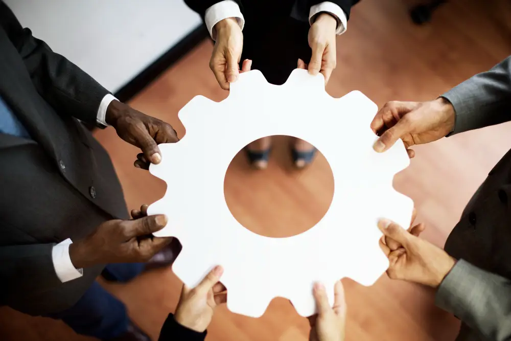 Fünf Personen im Business-Look halten gemeinsam ein grosses, weisses Zahnrad als Symbol für Arbeitssicherheit und Teamarbeit auf Holz.