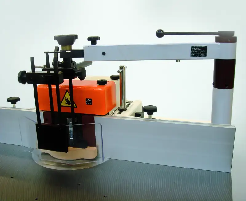 Detailansicht der Schwenkvorrichtung mit Arbeitsschutz-Haube, Einstellknöpfen und Metallteilen auf grauer Werkbank, Fuss 80 mm.