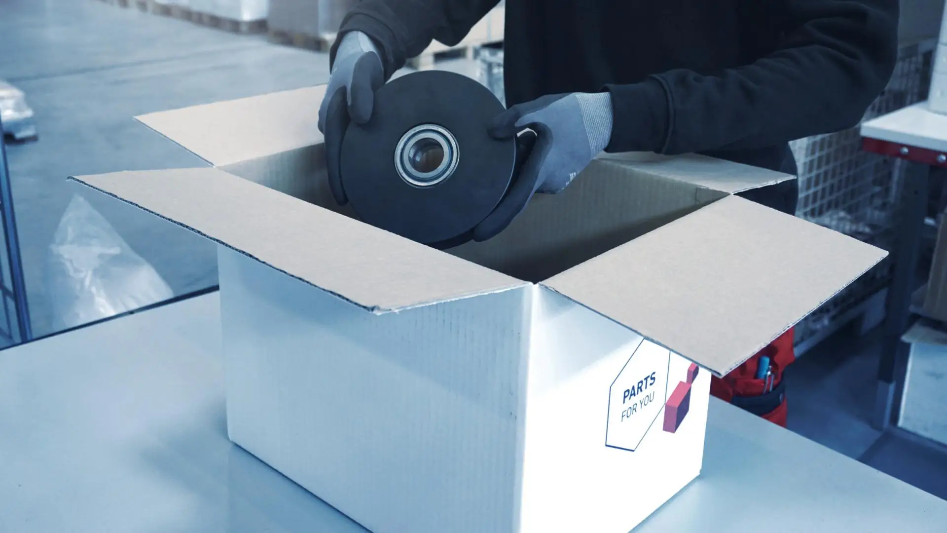 Eine Person mit Schutzhandschuhen legt Maschinenteil mit Kugellager in Karton "PARTS FOR YOU" im Lager, Arbeitsschutz garantiert.