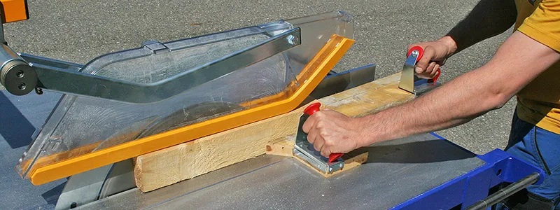 Eine Person führt Holz mit roten Schiebestöcken sicher durch die Tischkreissäge mit transparenter Schutzhaube.
