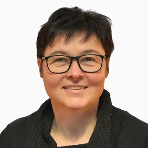 Person mit kurzen dunklen Haaren, Schutzbrille, lächelt. Schwarzes Oberteil mit Strukturkragen, Hintergrund ist weiß.